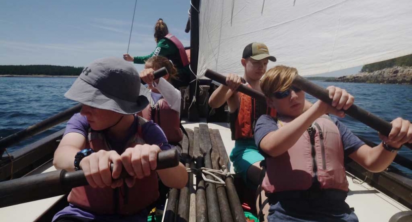 A group of students wearing lifejackets use oars to maneuver a sailboat. 